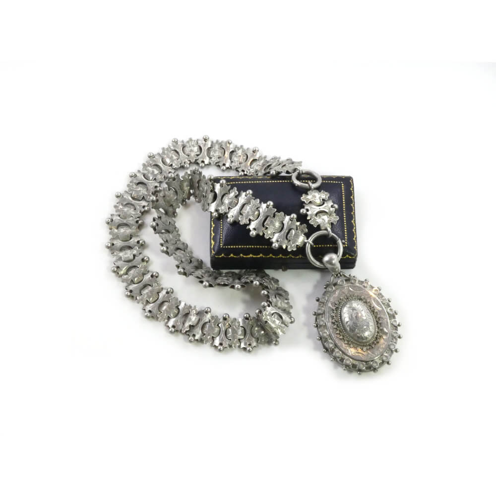 A Victorian Silver locket and chain displayed on a box, featuring intricately designed links and a large, ornate pendant with a textured surface and delicate floral decorative motifs by Hidden Gems.